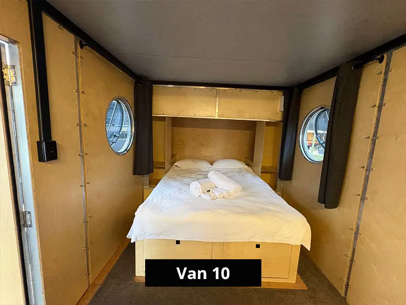 Cozy camper van bedroom with double bed, built-in storage, and round porthole windows in minimalist wooden interior – Van 10
