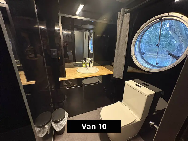 Modern camper van bathroom with toilet, round window, vanity sink, and mirror in a compact black interior design – Van 10