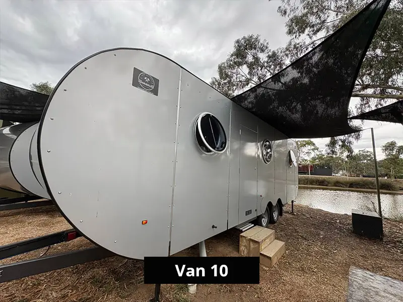 Modern compact camper van with rounded design, circular windows, and outdoor shade canopy parked beside a lake – Van 10 glamping accommodation