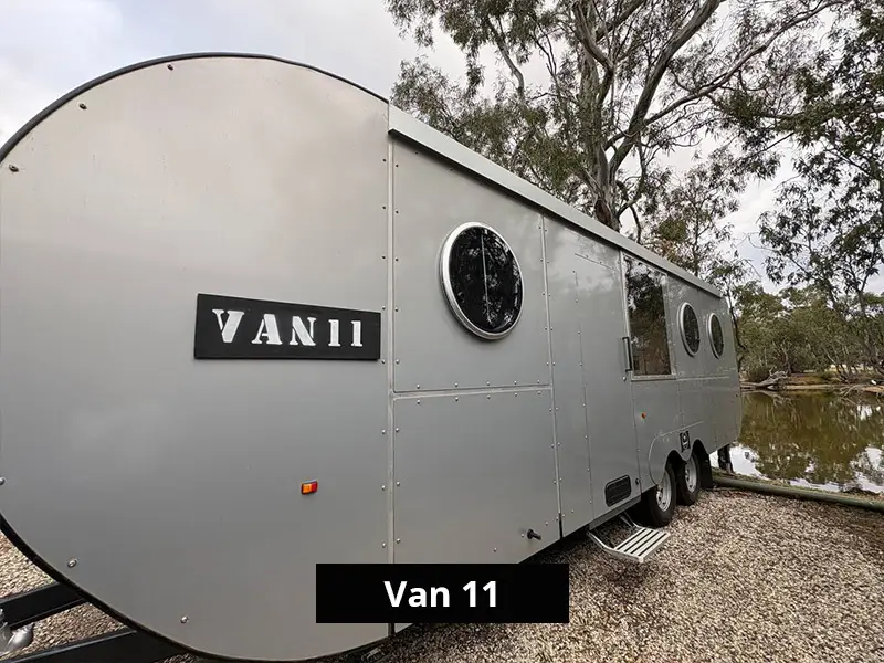 Modern grey camper trailer exterior labeled Van 11, parked by riverside with trees, featuring round windows and off-grid travel design