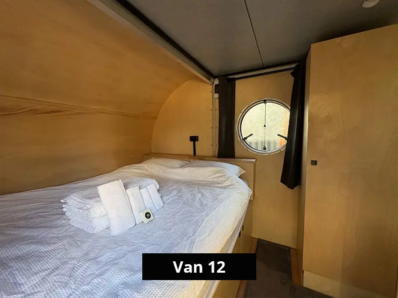 Retro van bedroom with double bed, white linens, wooden interior, and circular window, ideal for minimalist travel living