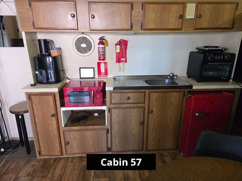 Compact cabin kitchenette with wooden cabinets, stainless steel sink, red microwave, mini retro-style refrigerator, coffee maker, and countertop oven in a cozy rustic cabin interior labeled Cabin 57.