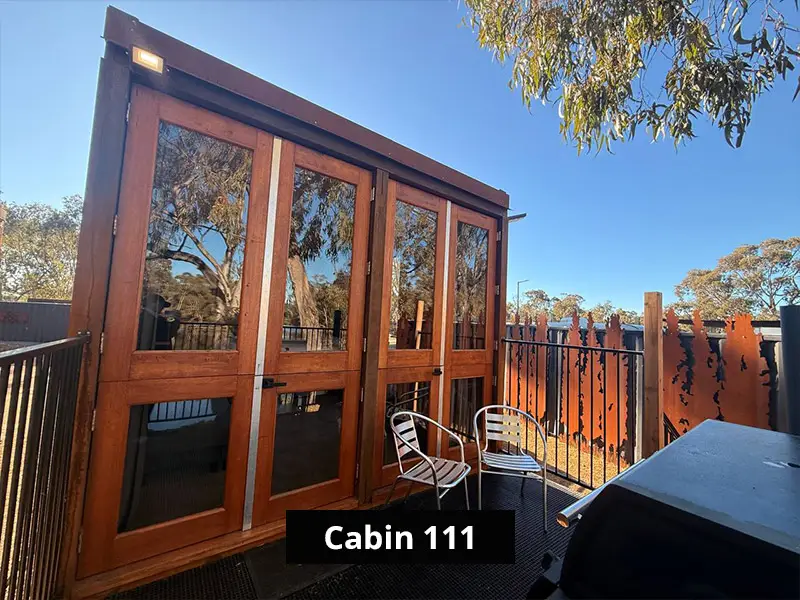 Cabin 111 exterior with timber-framed glass doors, private deck seating, barbecue area, and scenic bushland surroundings.