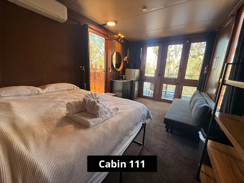 Cabin 111 interior with queen bed, folded towels, kitchenette, grey sofa, glass doors, and riverfront cabin views.