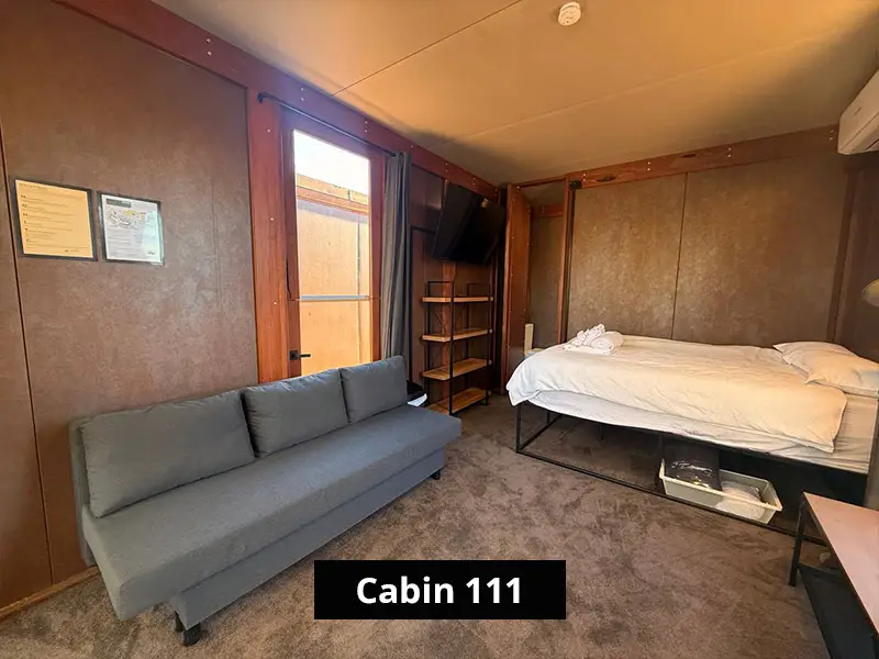 Modern Cabin 111 living space with grey sofa, white bed, wall-mounted TV, shelving, air conditioning, and warm brown walls.