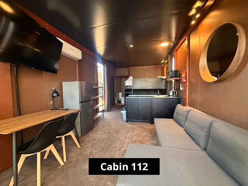 Modern Cabin 112 interior with grey sofa, kitchenette, bunk bed, refrigerator, wall-mounted TV, desk, and dining chairs.