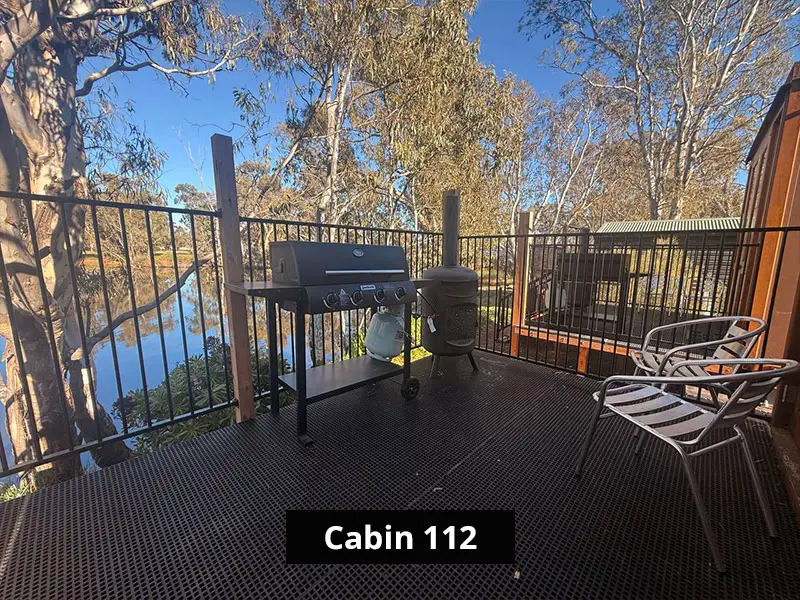 Private Cabin 112 outdoor deck with barbecue grill, metal chairs, black railing, and scenic riverfront bushland views.