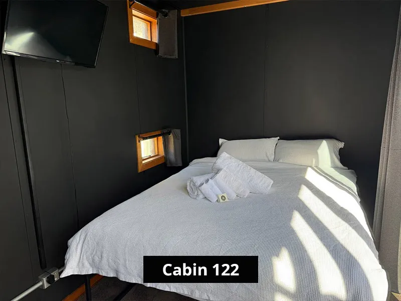Compact Cabin 122 bedroom with white double bed, fresh towels, dark walls, small windows, curtain, and wall-mounted TV.