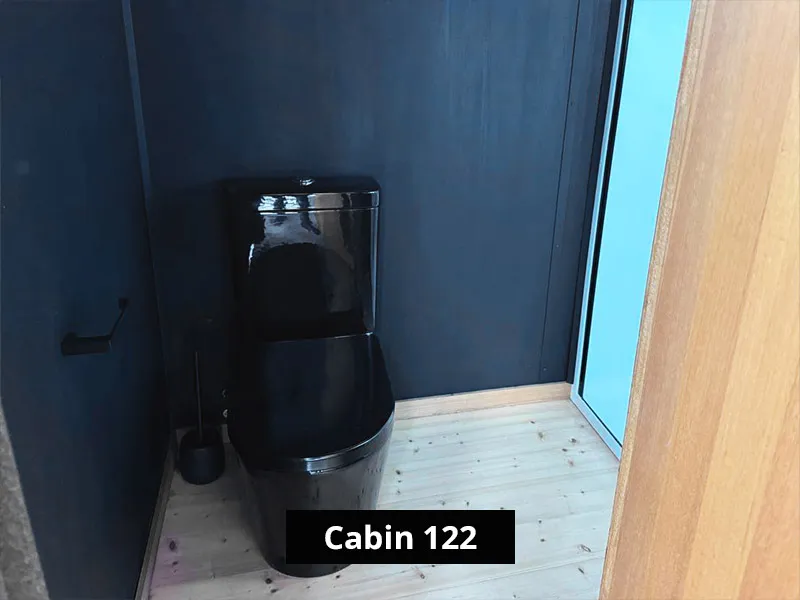 Cabin 122 bathroom toilet area with glossy black toilet, dark walls, black accessories, and light timber flooring.